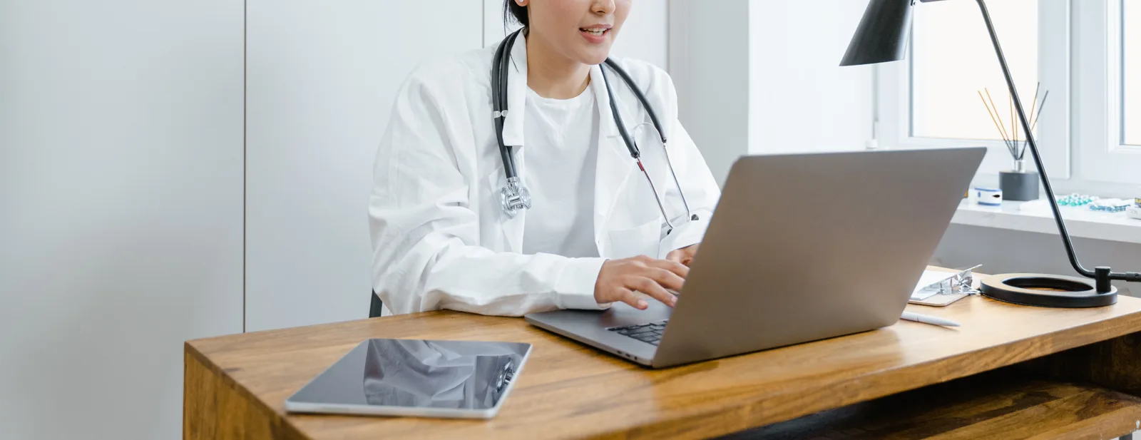 A clinician in a white coat working at a laptop in a bright office