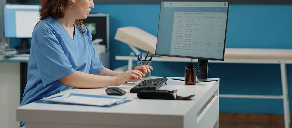 A nurse in scrubs working at a desktop computer in an exam room